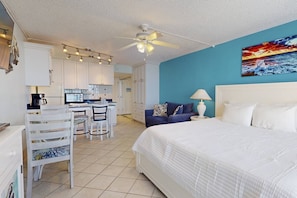 A hotel room with a bed, a desk with chairs, a kitchenette, and a ceiling fan.