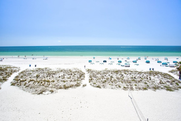 Une plage de sable blanc, avec une eau d’un bleu limpide et plusieurs parasols de plage bleus.