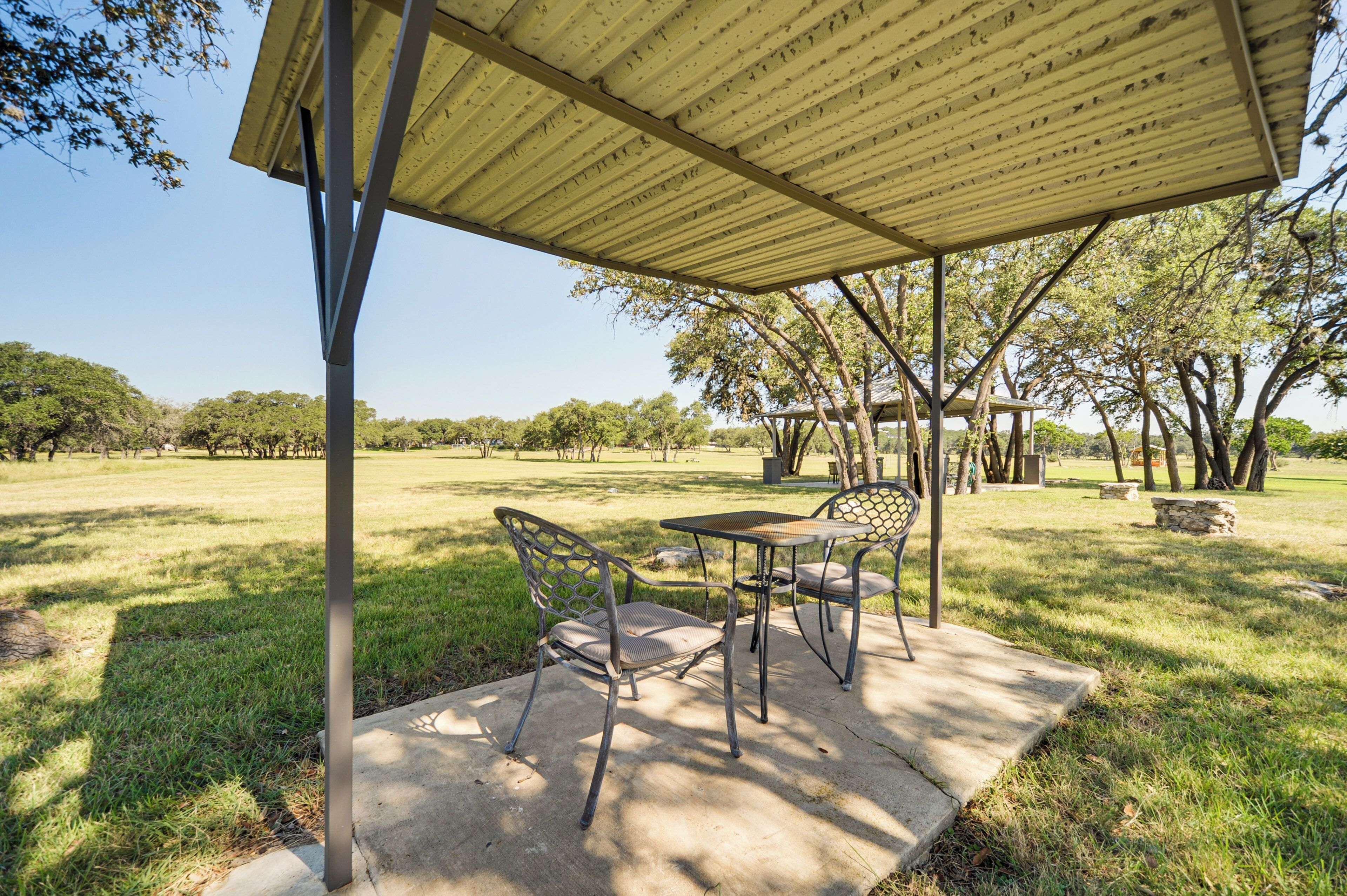 Un patio al aire libre con una mesa y sillas bajo un porche cubierto, rodeado de césped y árboles.