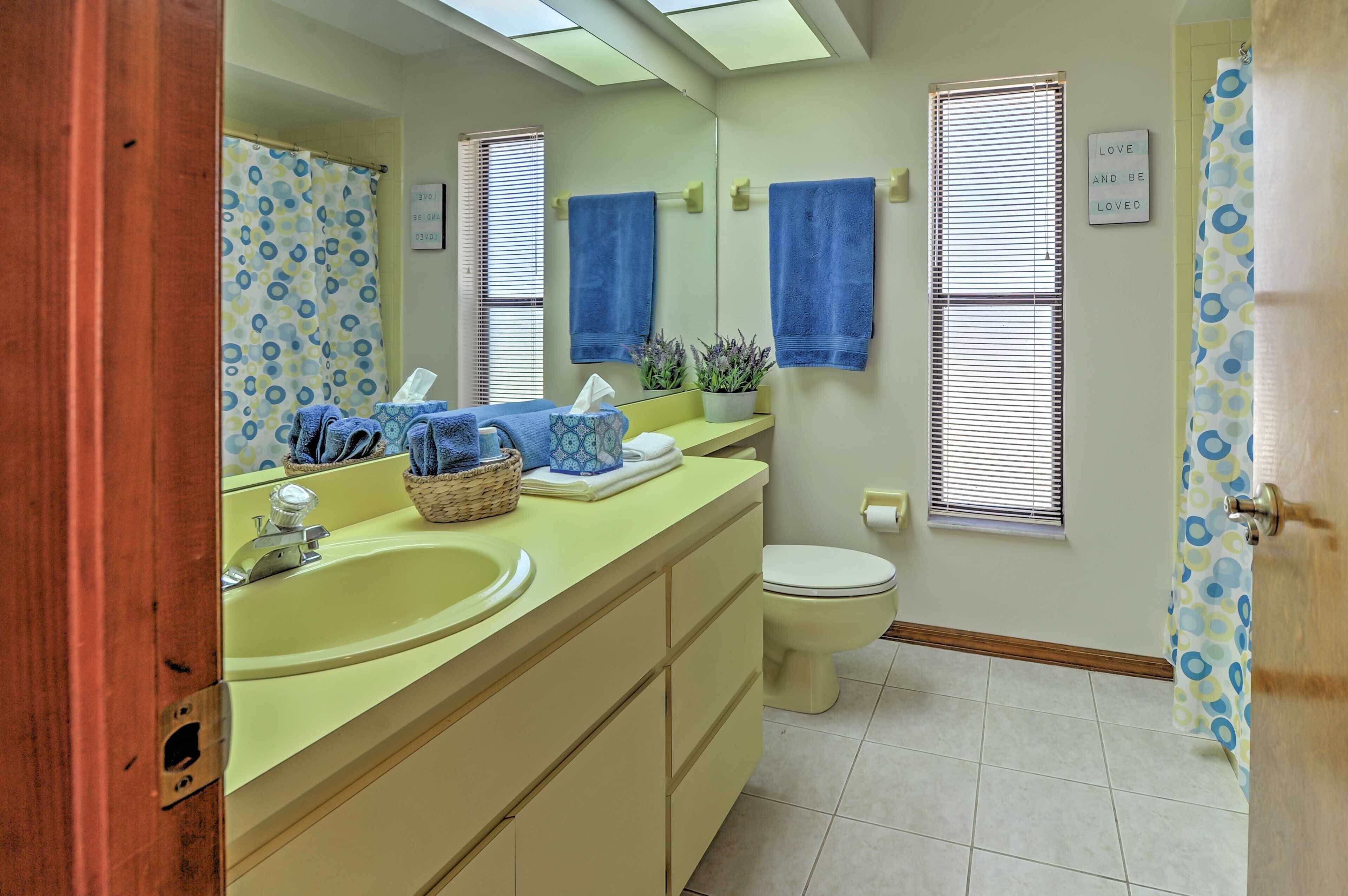 A bathroom with a yellow sink, a toilet, and a mirror. There are two towels hanging on the wall, and a window with blinds.