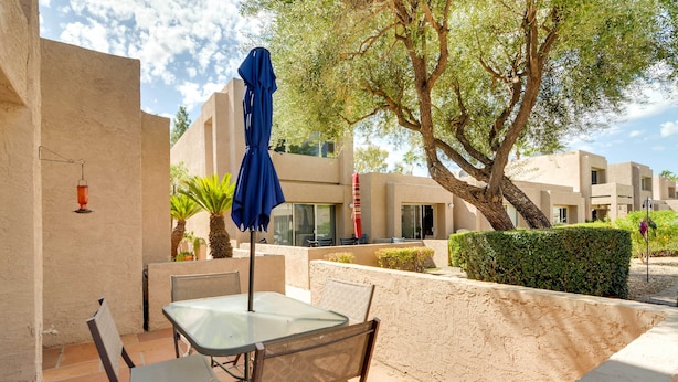 An outdoor patio with a glass table and chairs, a blue umbrella, and a large tree.