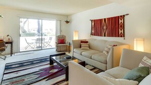 A living room with a sofa, armchairs, a coffee table, and a patterned rug. There is a sliding glass door leading to an outdoor area with a table and chairs.