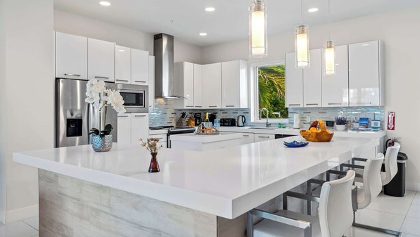 Una cocina moderna con armarios blancos, una isla grande, electrodomésticos de acero inoxidable, iluminación colgante y vistas al exterior con vegetación.