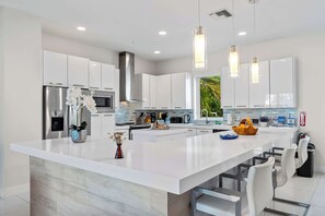 Une cuisine moderne avec des meubles blancs, un grand îlot central, des appareils en acier inoxydable, des luminaires suspendus et une vue sur la verdure à l’extérieur.