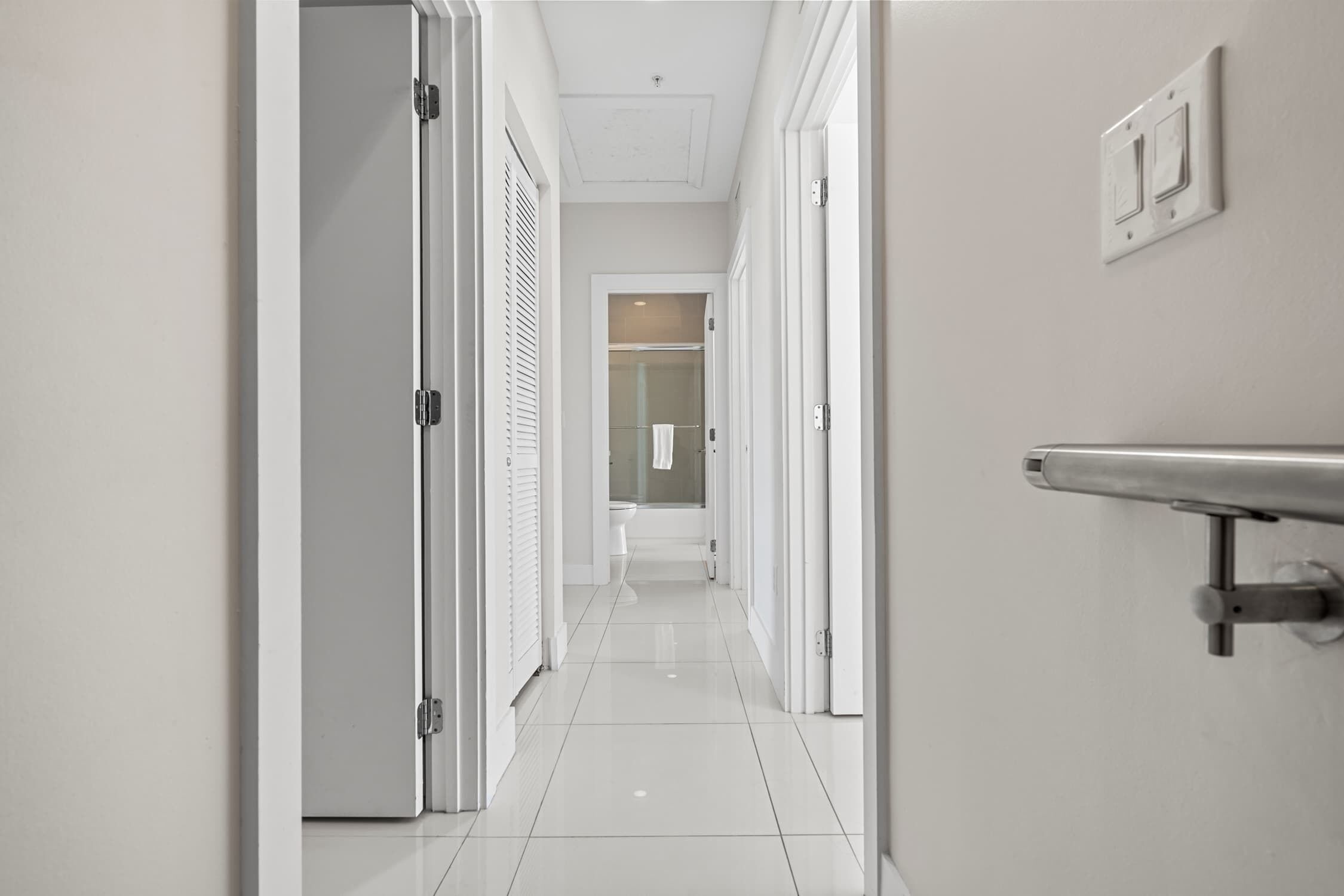 Moderni hotellikäytävä, jossa on valkoiset seinät, kiiltävä laattalattia sekä näkyvissä oleva kylpyhuone, jossa on wc ja suihku.