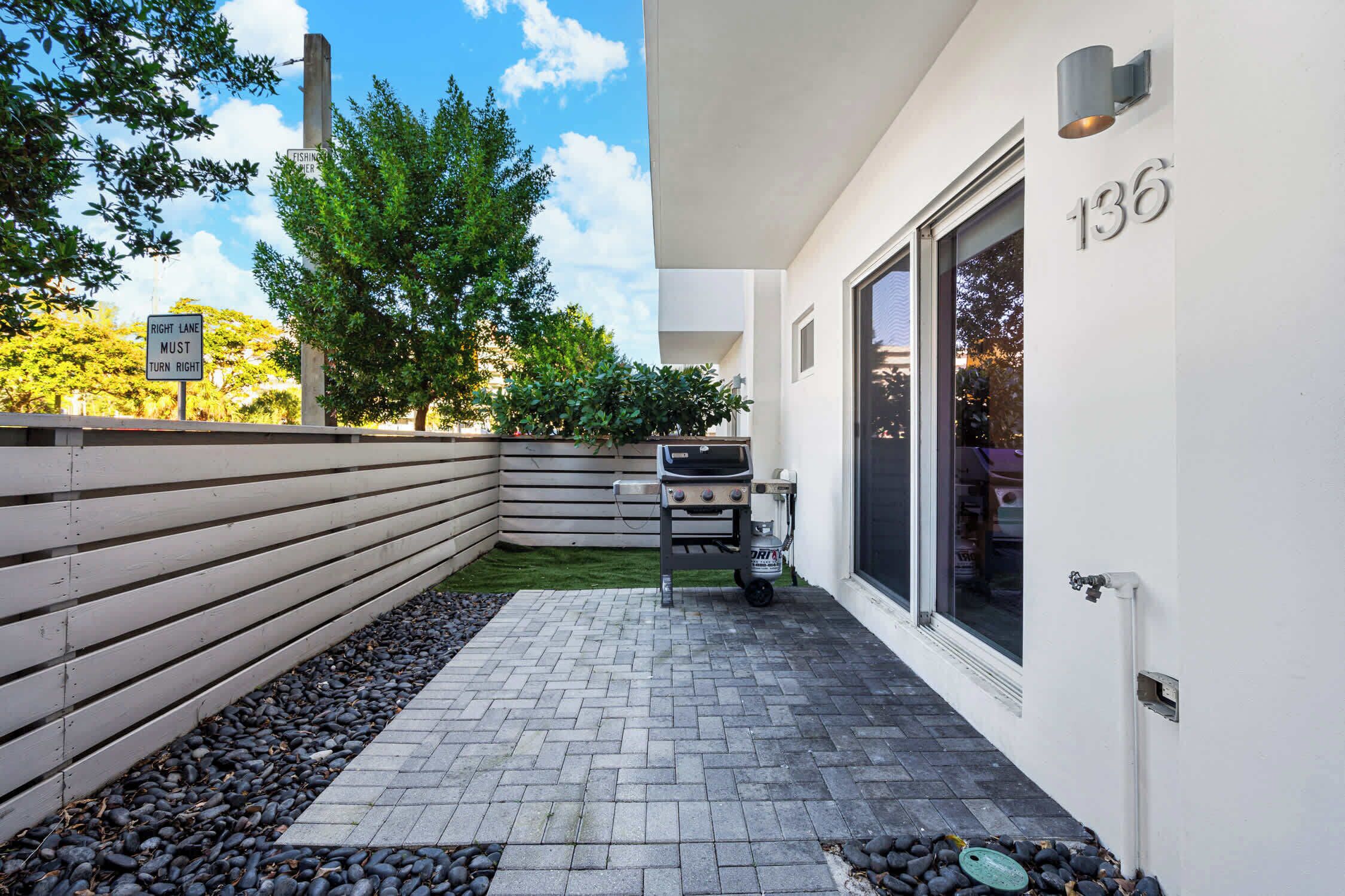 L’extérieur d’une maison moderne, avec une terrasse dallée, un barbecue et le numéro « 136 » peint sur le mur.