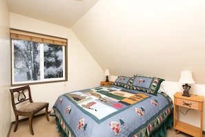 Quarto com cama grande, janela com vista para árvores, uma cadeira e mesa de cabeceira com abajur.