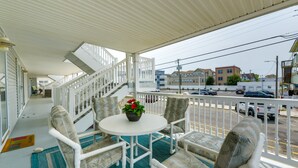 A covered outdoor seating area with white furniture and a table, overlooking a street with parked cars and buildings. - Condo w/ Deck: Walk to Beach & Convention Center! (Wildwood)