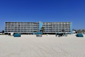 A large, multi-story hotel with numerous balconies, a sandy beach in front, and a clear blue sky.