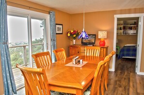 A dining area with a wooden table and chairs, a television, a vase with flowers, and a bookshelf in the background. - Ocean Grove Ocean View (Waldport)