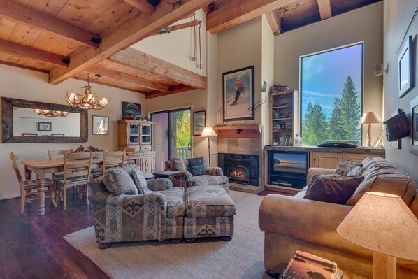A spacious living room with a fireplace, wooden beams, and a large window overlooking a forest.