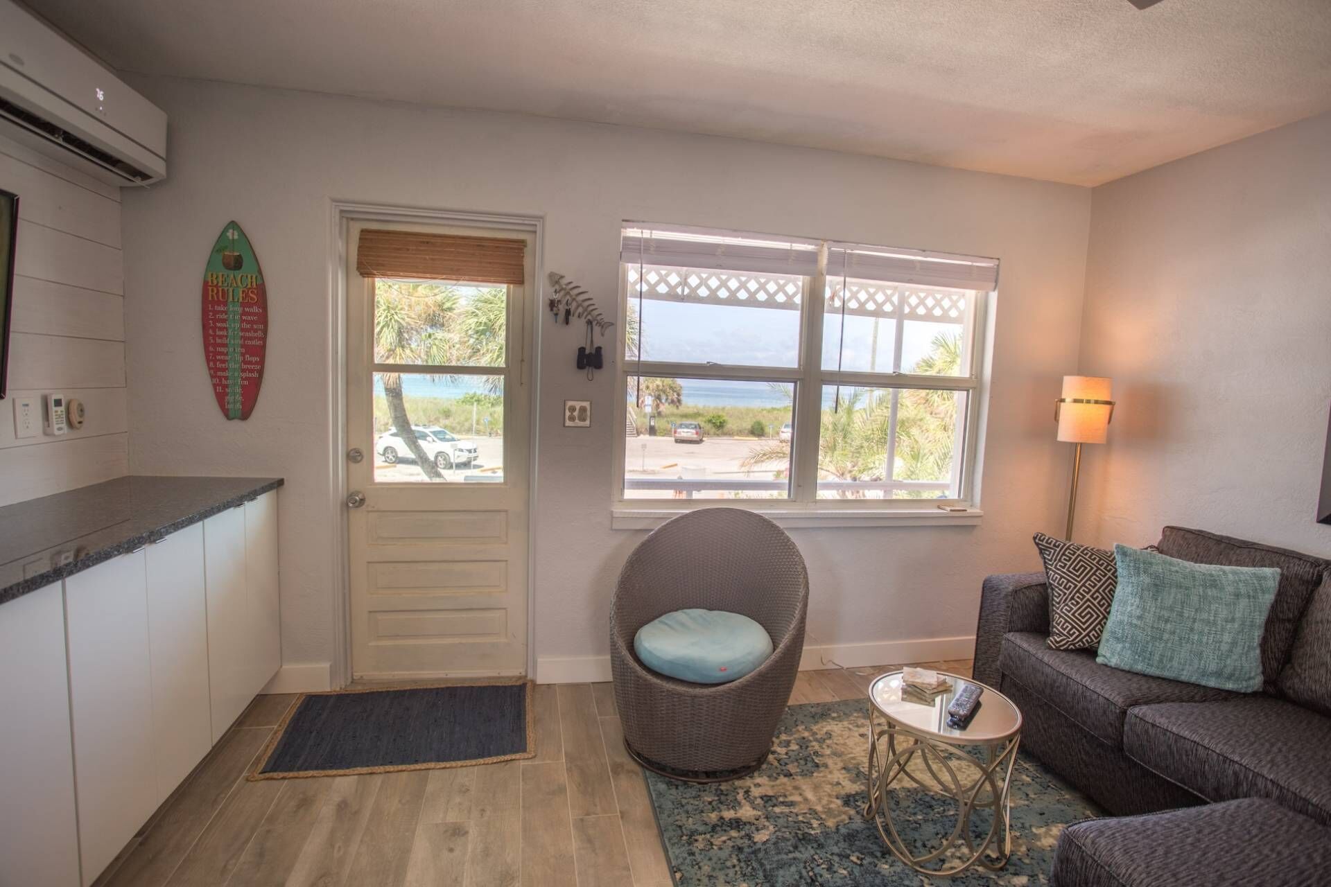 A compact living space with a kitchenette, a large window with a view of the beach, a wicker chair, and a sofa with cushions.