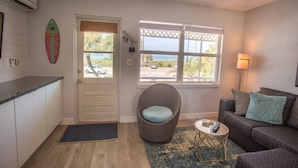 Un espacio habitable compacto con kitchenette, un ventanal con vista a la playa, una silla de mimbre y un sofá con cojines.