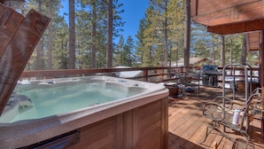 A wooden deck with a hot tub, outdoor seating, and a grill area surrounded by tall pine trees.