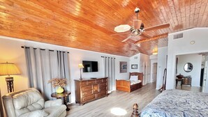 A spacious bedroom with a wooden ceiling, a large bed with a floral bedspread, a wooden dresser, a television, and a seating area with a chair and a lamp. - Playa del Sol: CL Lakefront + 3 Master Suites + Spacious Deck & Yard + Pool Table + Easy Lake Access (Canyon Lake)