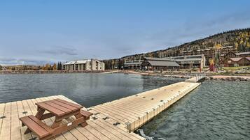 A wooden dock with a picnic table extending into a calm lake, surrounded by buildings with a mountainous backdrop.