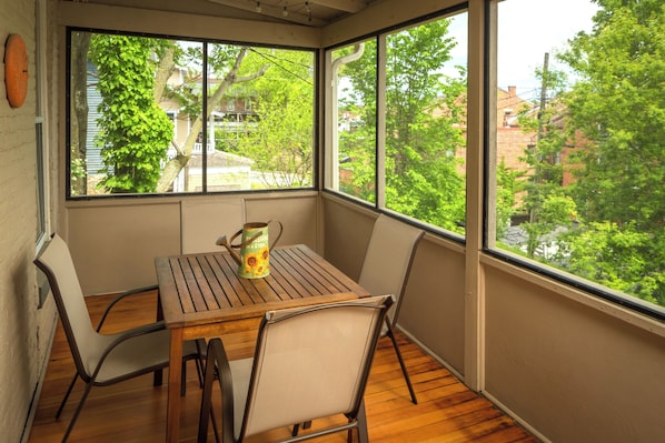 A sunlit room with a wooden table and chairs, a large window with a view of trees and buildings, and a decorative vase on the table.