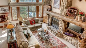 A cozy living room with a stone fireplace, a large window with a view of trees, a plush sofa, and a coffee table with decorative items.