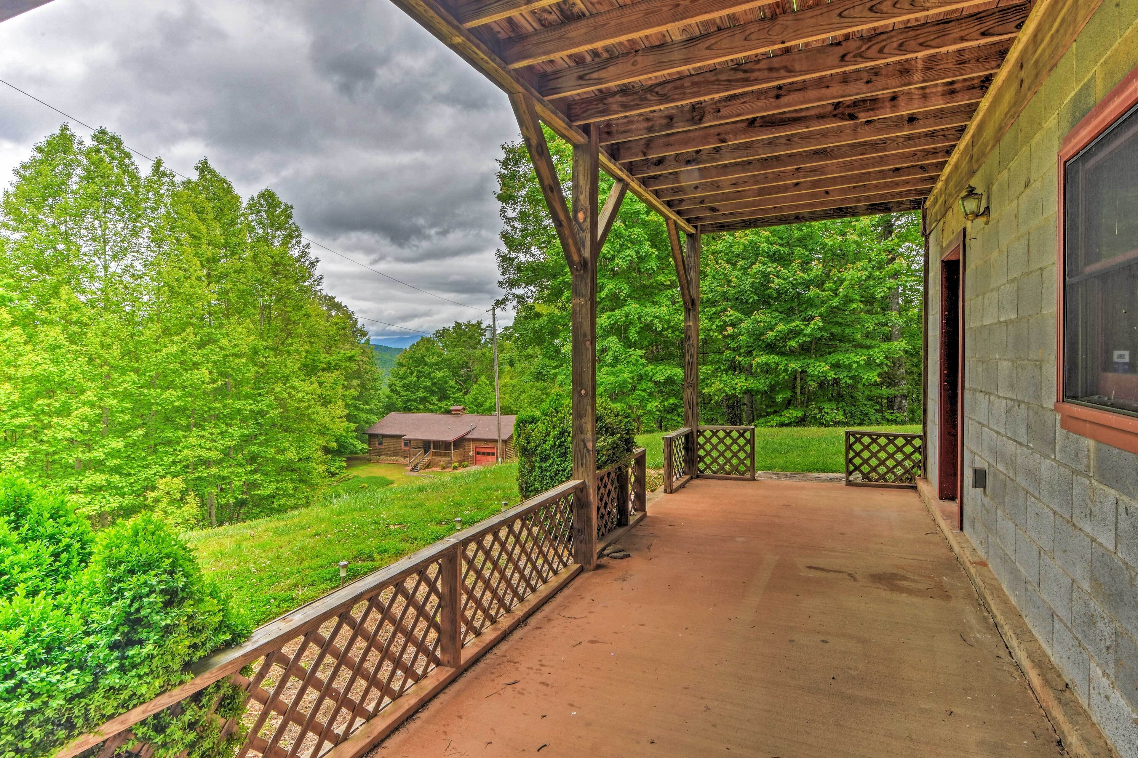 Une véranda couverte avec une balustrade en bois, un sol en béton et une vue sur un paysage verdoyant, avec des arbres et un bâtiment situé au loin.