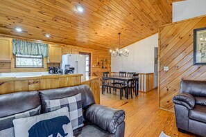 Un accogliente interno di una baita con soffitto in legno, zona pranzo, cucina con frigorifero e divano in pelle con un cuscino a forma d'orso.