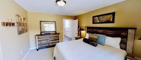 Quarto com uma cama grande, penteadeira, espelho e porta que dá para outro cômodo.