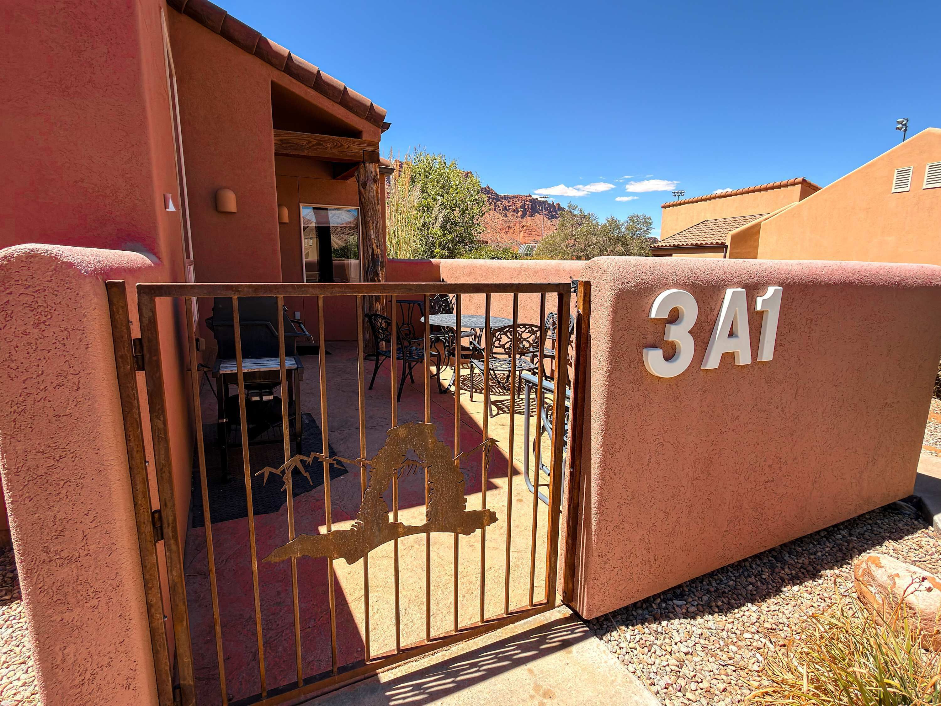 Una entrada protegida por un portón metálico con diseño decorativo, un patio con mobiliario exterior y un edificio con fachada de estuco y un letrero con el número '3A1'.