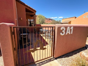 Una entrada protegida por un portón metálico con diseño decorativo, un patio con mobiliario exterior y un edificio con fachada de estuco y un letrero con el número '3A1'.