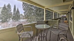 Ein überdachter Balkon mit Korbmöbeln und einem Glastisch, mit Blick auf eine verschneite Landschaft mit hohen Bäumen und Gebäuden.