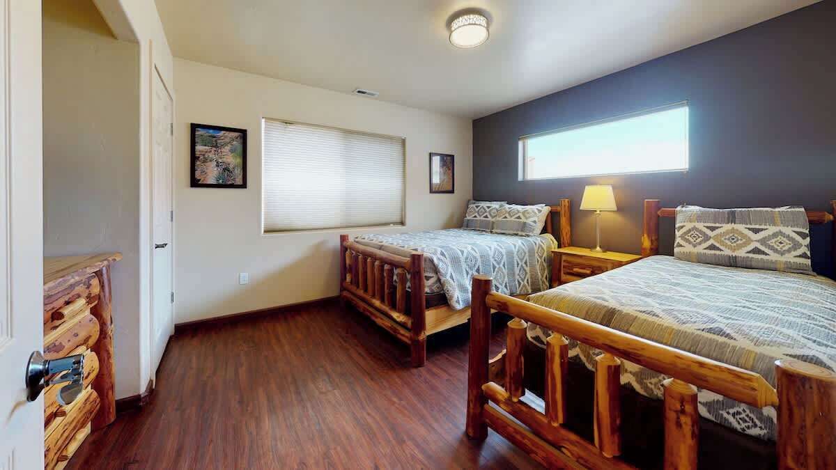 A bedroom with two wooden beds, a window with blinds, a nightstand with a lamp, and a door with a wooden handle.