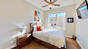 Une chambre à coucher comprenant un lit, une table de chevet, une lampe, un ventilateur de plafond, une fenêtre avec vue et un téléviseur fixé au mur.