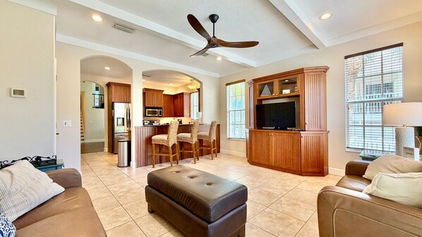 A spacious living room with a large TV, a ceiling fan, and a kitchen area in the background.