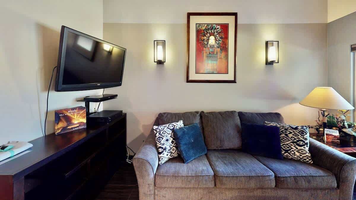 Ein Wohnzimmer mit Flachbildfernseher, einer gepolsterten Couch mit gemusterten Kissen, einem gerahmten Gemälde an der Wand und einer Stehlampe.