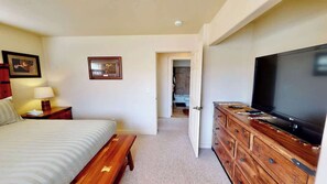 A hotel room with a bed, bedside table, lamp, dresser, television, and a door leading to a bathroom.