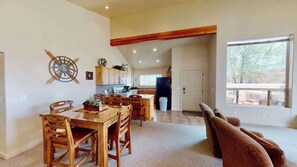 A spacious living area with a dining table and chairs, a kitchen in the background, and a large window providing natural light.
