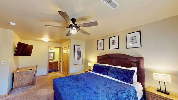 Un dormitorio con cama, ventilador de techo, escritorio y cuadros en la pared.