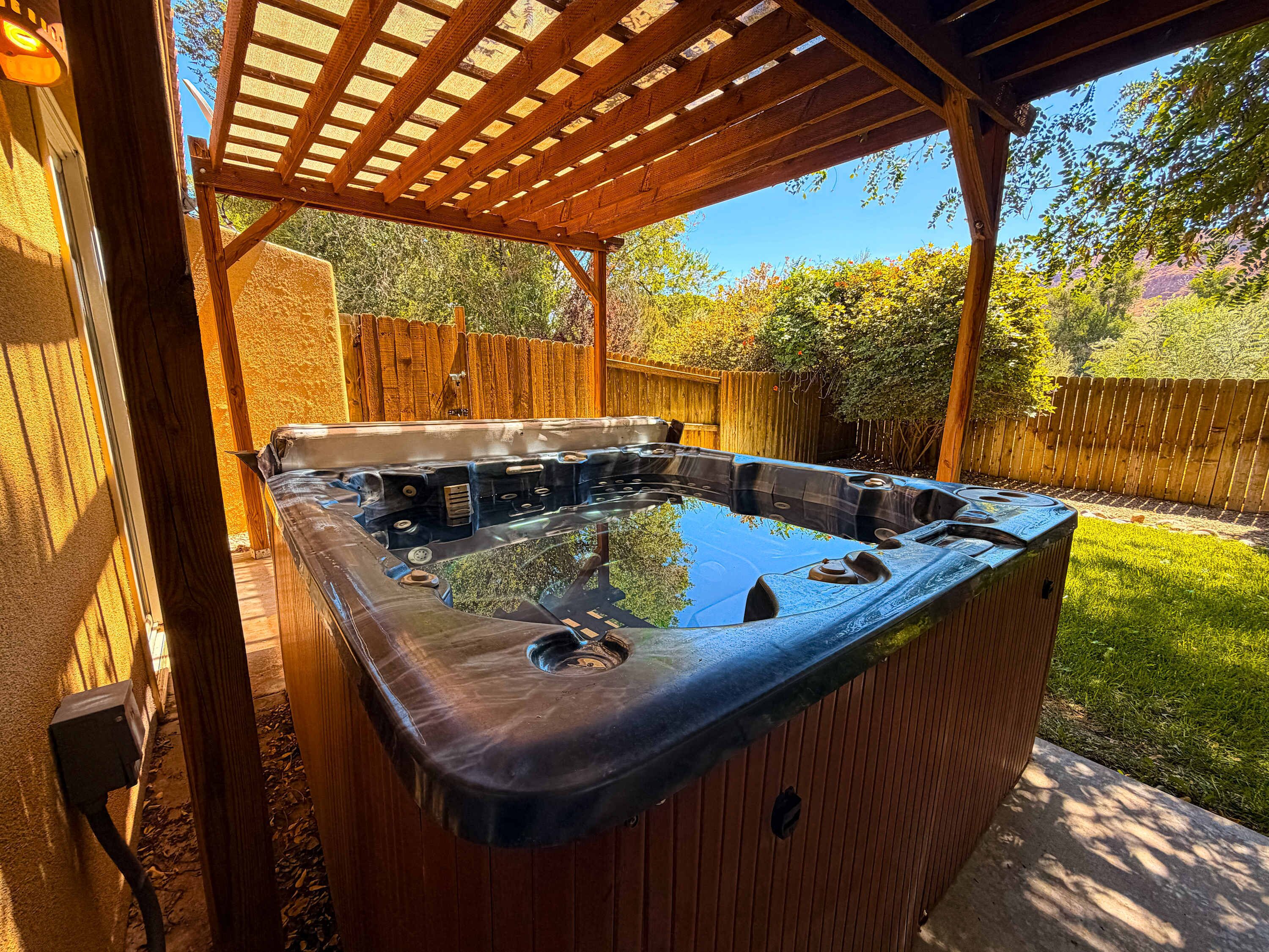 Un bain à remous sous une pergola en bois, dans un jardin avec une clôture en bois et de la verdure.