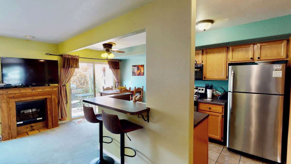 A kitchen with a refrigerator, wooden cabinets, a bar area with stools, and a television.