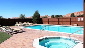 Piscina com espreguiçadeiras e banheira de hidromassagem, cercada por um muro de madeira e céu claro.
