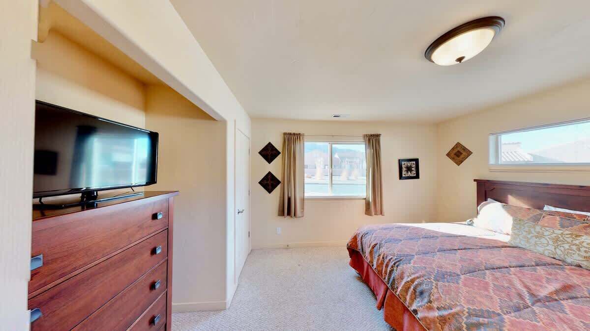 Une chambre à coucher comprenant un lit, une commode, un téléviseur, une fenêtre avec des rideaux et un luminaire au plafond.