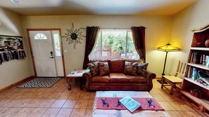 A living room with a brown leather sofa, a coffee table, a bookshelf, and a window with curtains. - Arches East - 389, Downtown Moab 3-Bedroom Townhom (Moab)