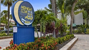 A sign for 'Midnight Cove' with a beach view, surrounded by tropical plants and palm trees. - Spacious vacation rental condo with lagoon views, beach access, and pool (Siesta Key)