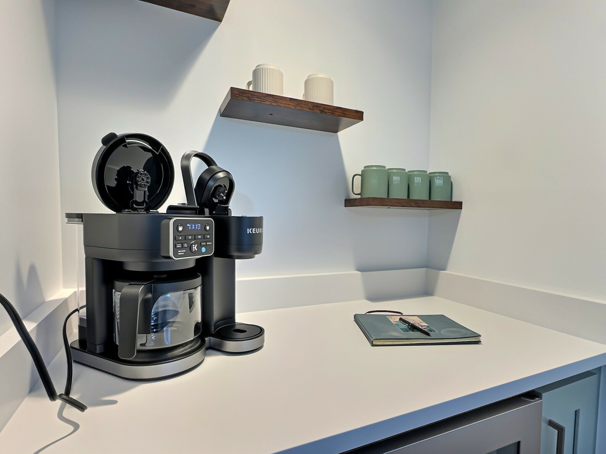 Una cafetera moderna sobre una encimera blanca, con una pantalla digital, un par de auriculares y un cuaderno y un bolígrafo.