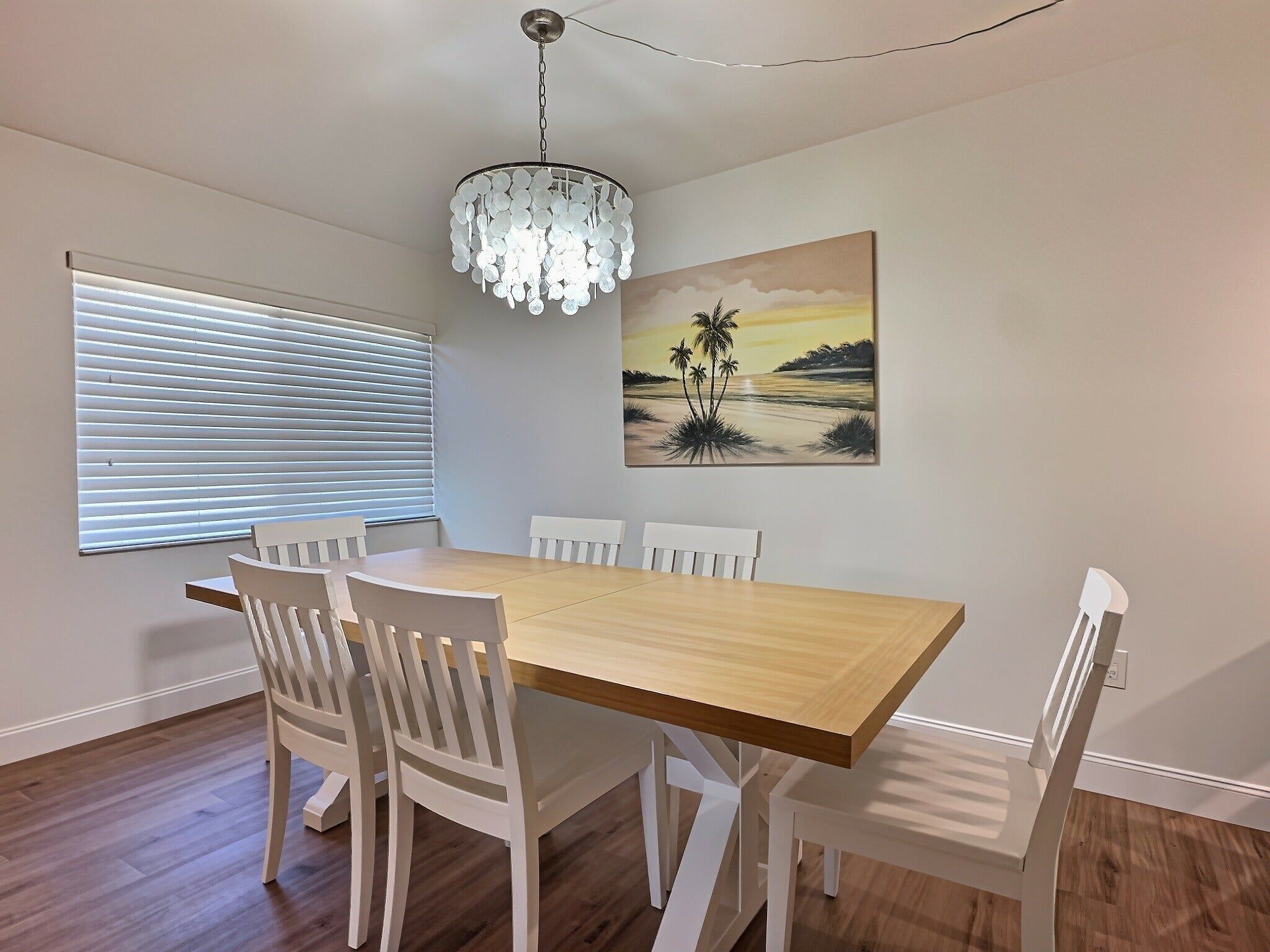 Un comedor con una mesa de madera y sillas blancas, un cuadro de una playa y una lámpara moderna.
