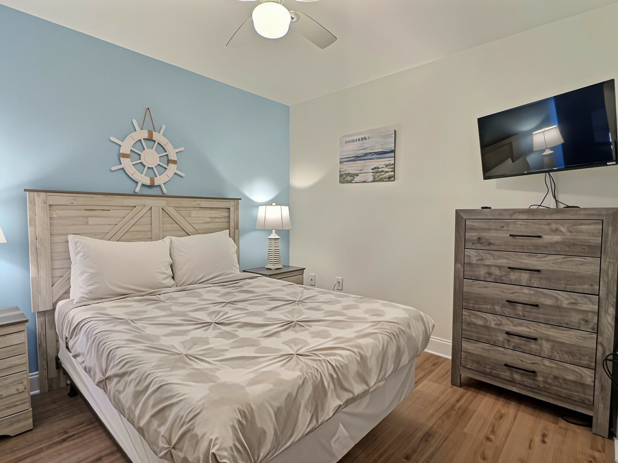 A bedroom with a bed, a nightstand, a lamp, a TV, a wooden dresser, and a wall-mounted ship's wheel decoration.