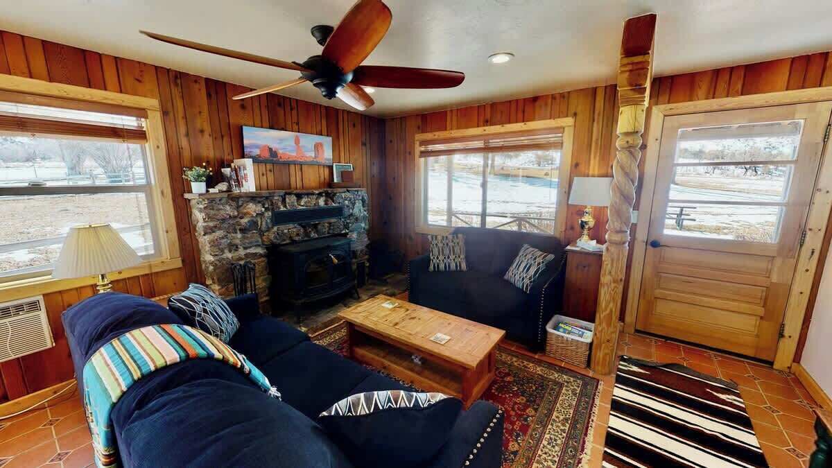 A cozy living room with a fireplace, wooden walls, a ceiling fan, and a large window.
