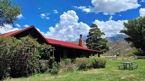 A rustic cabin with a red roof, surrounded by lush greenery and a clear blue sky.