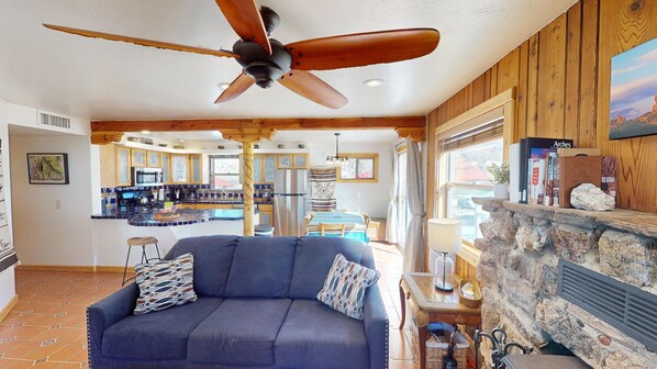 A spacious living area with a dark blue sofa, a wooden ceiling fan, a stone fireplace, and a kitchen in the background.