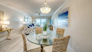 A modern living room with a glass-top dining table surrounded by wicker chairs, a chandelier, and a view of the ocean through large windows. - Vacation Villas #333 Gorgeous Beachfront Condo (Fort Myers Beach)
