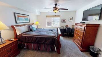 A bedroom with a bed, dresser, desk, chair, ceiling fan, window, and television.