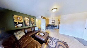 A living room with a brown leather sofa, a coffee table, and wall art. There is a dining area with a table and chairs in the background.